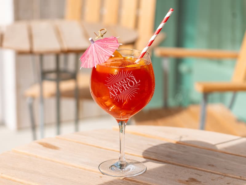 Stort stilkglas med graveret tekst, fyldt med en orange drink med sugerør og lyserød parasol, på et træbord udenfor